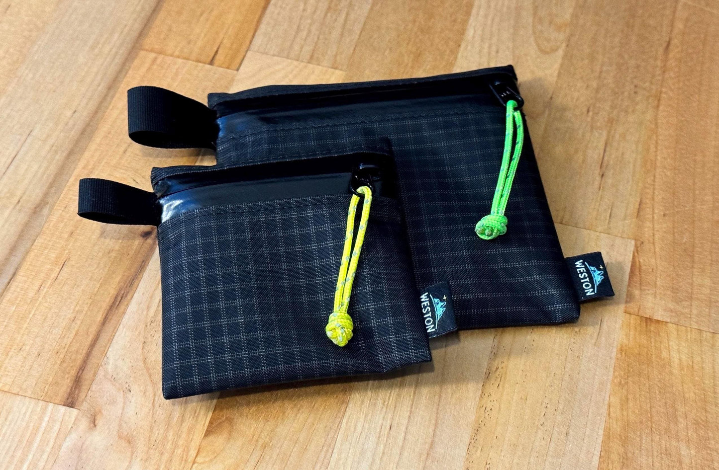 May include: Two black grid-patterned zipper pouches with black straps and bright yellow and green pull cords.  The smaller pouch is on top of the larger pouch. Both pouches have a Weston logo label. These are lightweight, durable, and compact storage solutions ideal for travel or organization.