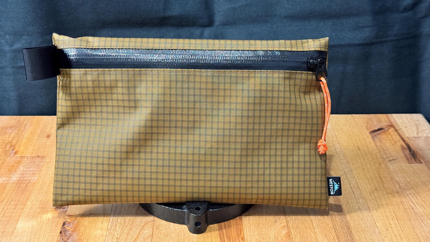May include: A tan and brown grid patterned zipper pouch with a black zipper and an orange pull tab.  The pouch has a black strap on one side and a small woven label is visible on the bottom right corner. The pouch is displayed on a wooden surface.  This is a lightweight, durable, and versatile zipper pouch ideal for everyday carry or travel.